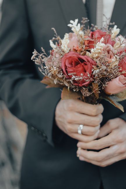 A person dressed in a dark suit is holding a bouquet of fresh flowers, featuring large, softly pink and coral roses, complemented by delicate sprigs of white and lavender-hued filler flowers. The rose