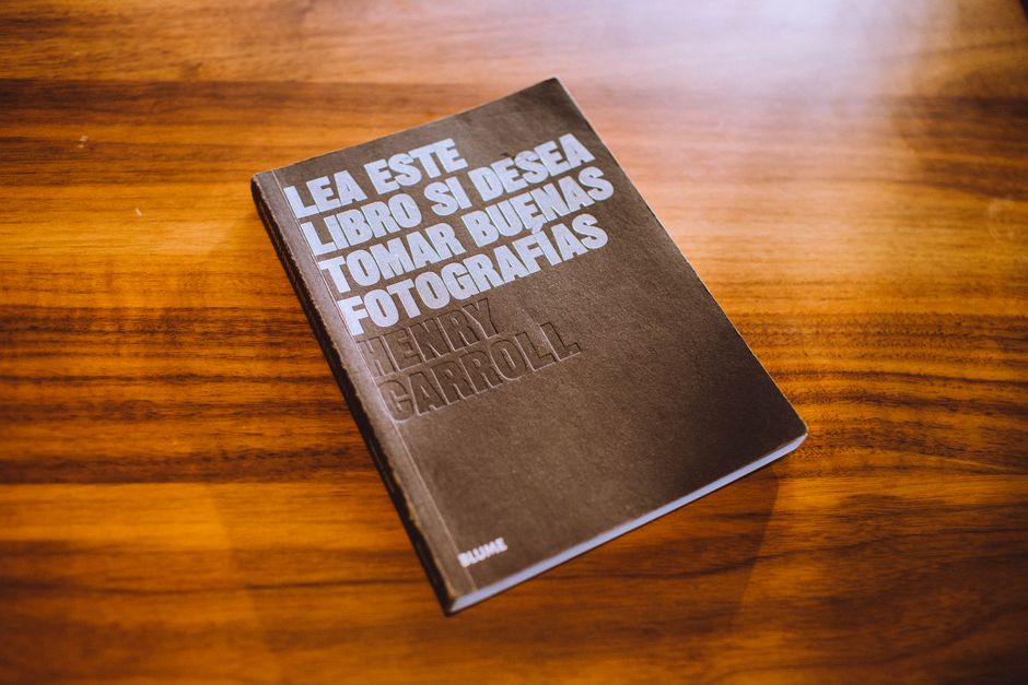 A black hardcover book titled 'Lea Este Libro Si Desea Tomar Buenas Fotografías' by Henry Carroll rests on a polished wooden surface with a warm, orange-brown tone. The book's title is embossed in la