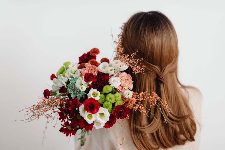 A woman with long, wavy auburn hair, styled with a small braid on one side, holds a large, vibrant bouquet of fresh flowers in front of her shoulder. The bouquet features a mix of red, white, pink, an