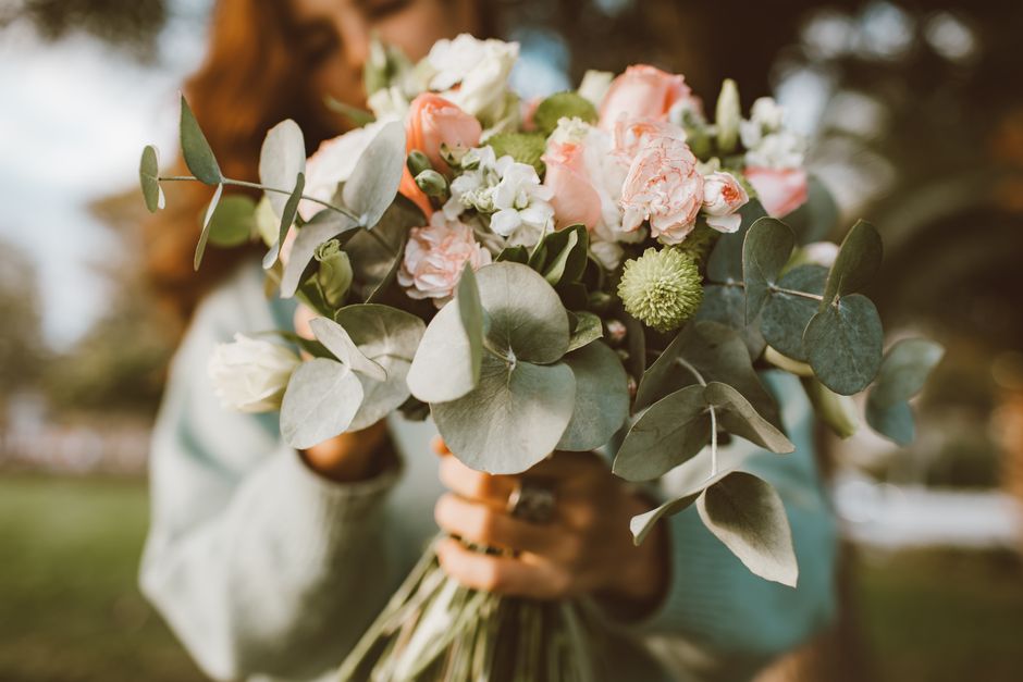A close-up of a floral bouquet held by a person, featuring a mix of soft pink and white roses, delicate white lisianthus, and round green scabiosa flowers, all accented with fresh, silvery eucalyptus