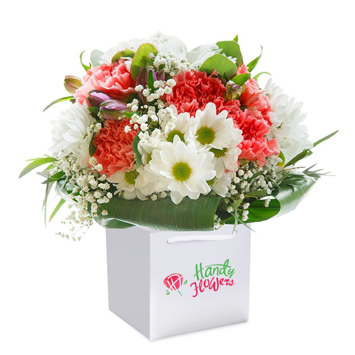 Set against a flawless white backdrop, this floral bouquet is arranged upright in a sleek square gift bag with shimmering ribbon handles-a familiar sight among Lea Bridge's flower lovers. The heart of the composition brims with coral-pink and deep red carnations, each petal curled in intricate ruffles, glowing with warmth and energy. Threaded between these stars, snowy white daisies with luminous green-yellow centers add lively contrast, conjuring the hopeful mood of spring mornings along Millfields Park. Scattered sprigs of baby's breath lend an airy, innocent touch, their tiny blossoms lighting up the arrangement like dew. Deep green leaves arc around the bouquet's foundation, their shine and shape providing elegant structure and lushness-an ode to the leafy avenues of Walthamstow and Hackney Marshes nearby. Small, purple-tinged closed buds nestle discreetly on one side, hinting at the unfolding beauty just beneath the surface. The entire piece is balanced in a rounded, abundant form that radiates joyous abundance, while the understated white gift bag emphasizes ease of delivery-a nod to the practicality locals value. This detailed bouquet embodies the thoughtful craftsmanship of Lea Bridge's florists, ideal for birthdays, heartfelt celebrations, or simply to share a moment of beauty within the vibrant E10 community.