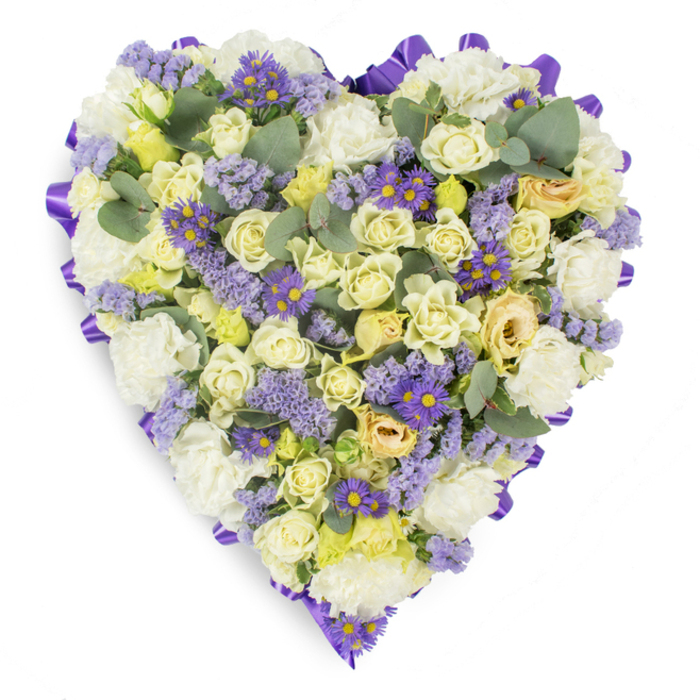 Seen against a pristine white background, this heart-shaped sympathy wreath is a study in gentle contrasts and careful craftsmanship: a dense, hand-placed field of ivory and pale yellow roses creates a soft, luminous centre, their petals layered like folded silk to form comforting depth. Nestled among them, pristine white carnations add rounded volume and a classic, consoling presence, while small sprays of lavender statice and vivid purple aster-like flowers with bright yellow centres thread through the arrangement, introducing tender flashes of cool colour and a textured, meadow-like detail. Sprigs of blue-green eucalyptus are woven thoughtfully throughout, their muted, rounded leaves offering visual pauses and an implied, fresh herbal scent that relaxes the composition. The heart's outline is crisply defined by a continuous, looped purple satin ribbon that acts as a refined frame, its sheen catching the light and giving the tribute a formal, finished feel suitable for funeral chapels, memorial services, or a graveside dedication. The overall palette-soft whites, creams, pale yellows, and lavender tones-evokes peaceful remembrance and enduring affection. As you might expect from a neighbourhood florist mindful of local rituals, this piece would sit respectfully at a service near Lea Bridge station or beside a family plot after a quiet procession along the River Lea, offering a quiet, dignified expression of sympathy and love.