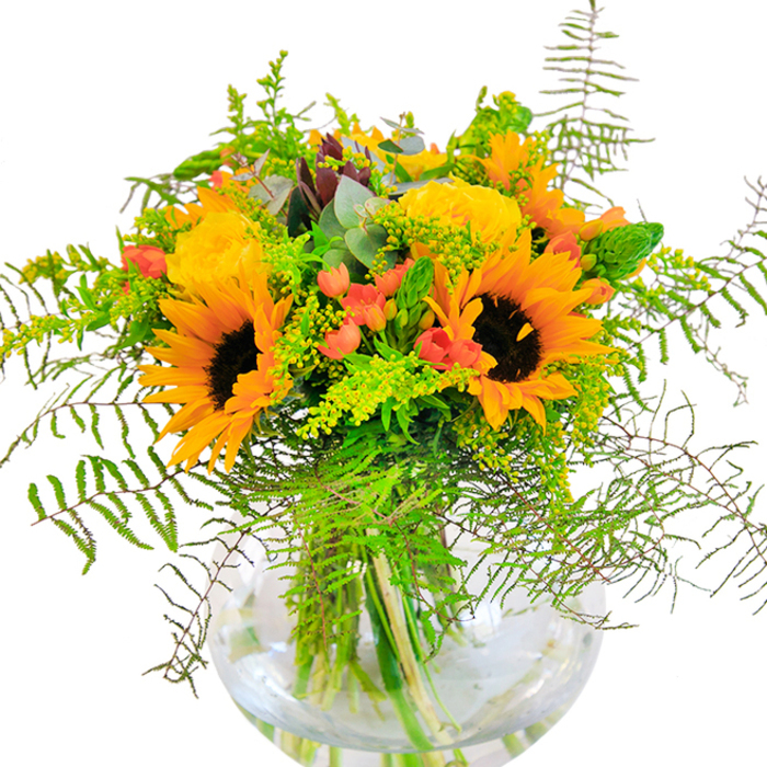 The first sensation this bouquet brings is one of luminous cheer, reminiscent of sunlit mornings near Lea Bridge's bustling junctions. Bold sunflowers, their petals aglow in golden yellow gradients, stand tall beside layers of plush yellow garden roses, each a marvel of delicately spiralled form. Deep green ferns tumble outward, softening the border of the arrangement, their light touch evoking the wildness of local green corridors. Between the blooms, clusters of glossy red Hypericum berries glisten, their colour playfully contrasting against the muted tones of eucalyptus and solidago. The entire display is arranged in a rounded glass vase, where the intricate network of stems is on full view-submerged in clear water to preserve their freshness through the bustle and calm of daily life in Lea Bridge. Viewed up close, the textures dance: the velvety faces of sunflowers, the silk-like petals of roses, the crisp movement of foliage. This arrangement would grace a table or countertop for any joyful occasion, its scent and colour recalling the bright promise of the community's summer festivals and riverside gatherings. The bouquet is a testament to the skill and passion of local florists who infuse every creation with heart and artistry.
