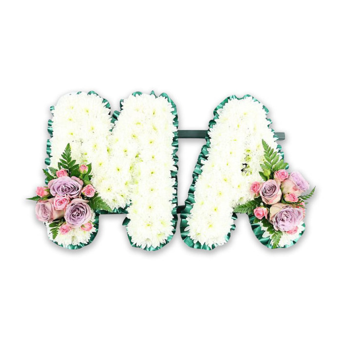 A personalised funeral tribute in the form of the letters MA is presented here with striking clarity, the design simple yet full of emotion. Both letters are built on sturdy frames and lie flat against a bright white background, giving the arrangement a clean, uncluttered look as if it were displayed on a studio table in a Lea Bridge flower shop. The florist has covered every part of the M and the A with compact white chrysanthemums, placed so closely that almost no gaps can be seen. The flowers form a continuous, cloud-like surface, each bloom with a faint green centre and feathery petals radiating outward, conveying purity and peace. Tracing the outline of each letter is a fold of deep green foliage or ribboned greenery, crisply pleated along the edges, which provides a strong visual boundary and adds a polished, tailored feel. This dark green border echoes the natural tones you might notice in Millfields Park on a quiet afternoon. At the base of each letter, two symmetrical arrangements sit like small corsages. On the left foot of the M, and the right foot of the A, clusters of pastel roses unfold in layers of soft colour: blush pink roses, cool lilac roses, and dainty pink spray roses are nestled together, with a few buds waiting to open. Delicate fern leaves and sprigs of foliage stretch gently outwards, breaking the straight lines and bringing a sense of organic movement. The overall mood is gentle, thoughtful, and deeply respectful, ideal for a family in Lea Bridge wishing to honour a cherished mother or Mama with a calm, elegant floral message at a funeral or memorial gathering.