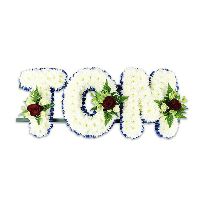 A carefully composed funeral tribute that spells TOM in bold, voluminous letters, each letter entirely covered with countless small, immaculate white chrysanthemum blooms to create a plush, tactile surface. The edges of every letter are neatly finished with a vivid royal blue ribbon, its smooth sheen drawing a refined border that sharpens the letterforms against the pristine white background. Within the central area of each letter sits a restrained floral spray: a single deep, scarlet-red rose forms the visual and emotional focal point, surrounded by smaller creamy white spray roses and graceful fern fronds which introduce textural contrast and verdant lines. The letters are supported by a slim dark green or black metal framework and a faint horizontal bar visible behind the base, a discreet structural element that ensures stability when placed on an easel or at a graveside. The composition is lit in soft, even light so the white chrysanthemums glow with quiet dignity and the blue ribbon and red roses read with clarity. There is a gentle fragrance of roses tempered by the green scent of foliage. As a bespoke sympathy piece made by Flowers Lea Bridge, this design is intended for memorial services across the local area - from intimate chapel gatherings near Lea Bridge Road to committals close to Walthamstow Marshes - offering a personal and respectful way to honour a loved one with craftsmanship and care.