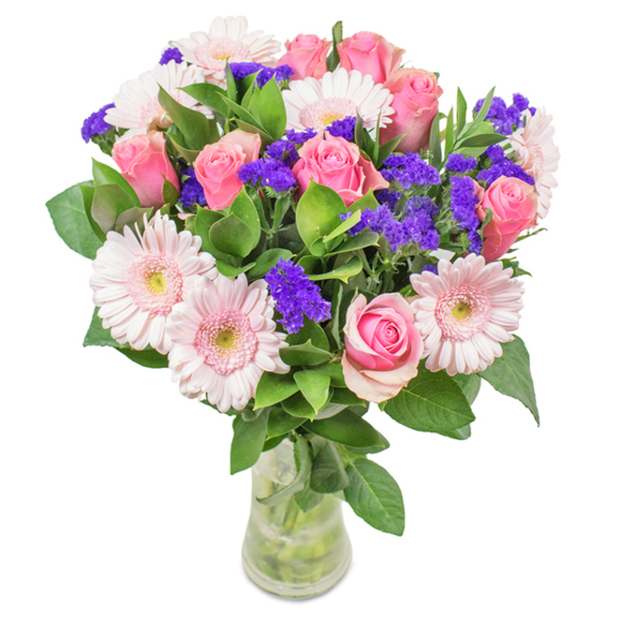 A radiant, meticulously arranged bouquet rests in a slender clear glass vase, poised against a pure white backdrop as if awaiting its role in a cherished Lea Bridge celebration. At its core, velvety pink roses-ranging from tightly furled buds to those just beginning to blossom-display the classic, spiral beauty for which they are beloved. Around them, a generous scattering of blush pink Gerbera daisies with large, soft heads and pale, almost luminescent centres draws the eye, inviting touch and admiration. The arrangement is enlivened by purposeful clusters of vivid purple statice, whose papery texture and rich colour provide striking contrast and visual depth, echoing hues found in local wildflower patches near Hackney Marshes. Ample deep green foliage fans out beneath the blooms, infusing the bouquet with a sense of lushness and vitality, like a pocket of spring thriving in the heart of Lea Bridge. The transparency of the vase allows the stems to be admired, and the clear water shimmers with freshness-a subtle promise of the care that has gone into each choice. This bouquet feels right at home in moments of celebration: as an anniversary centrepiece, a birthday surprise, or a meaningful way to say thank you or thinking of you within the lively bonds of the community. Elegance mingles with joy in every detail, making it a perfect gesture for any heartfelt occasion in Lea Bridge.