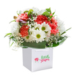 There is an unspoken joy in this bouquet, which captures the light, airy mood of a fresh Lea Bridge morning. Coral and deep red carnations, lush with gently ruffled petals, seem to celebrate right alongside the radiant white daisies whose golden-green centers bring a sense of cheerful optimism-much like the laughter heard along the edge of Jubilee Park. Wisps of white baby's breath create a whimsical, floating effect, scattered like confetti between the larger blooms. Subtle nods of unopened purple-tinged buds tuck among the clear, shining green leaves, adding a quiet sense of anticipation and richness to the design. The whole arrangement is neatly nestled within a modern white gift bag, whose ribbon handles catch the light, suggesting an easy journey from the florist's shop to someone's front door along Lea Bridge Road. Every detail feels intentional and full of promise-whether marking a birthday, an anniversary, or simply inviting a friend to smile. The freshness, abundance, and elegant artistry speak of local pride and a deep understanding of how flowers can brighten any corner of East London.