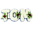 A personalised funeral flower arrangement spelling out the name TOM is displayed in three sturdy block capital letters, the kind of meaningful tribute often chosen by families in Lea Bridge for a final goodbye. Every letter is covered completely with closely packed white chrysanthemums, creating a smooth yet softly textured surface that reads clearly from a distance while still looking gentle and comforting up close. Bordering the outer edges of the letters is a rich royal blue satin ribbon, carefully pleated to follow each curve and corner, giving the piece a defined outline and a touch of formality. Resting on the letters at select points are small, artfully composed sprays of flowers: deep, velvety red roses as the emotional heart of each cluster, accompanied by petite white or cream spray roses and slim, trumpet-shaped white blooms with a freesia-like elegance. These are cushioned by pockets of lush green foliage-ferns and mixed leafy greens-that add depth, shape, and a suggestion of living garden growth. The letters sit side by side on a slim, dark rail, indicating a secure frame suitable for resting on a coffin top, hearse, or stand at a memorial service in a local church or crematorium serving Lea Bridge and nearby areas. Set against a plain white background, every hue and texture stands out in clear relief. The overall impression is of a bespoke sympathy tribute: dignified, tender, and unmistakably personal, created to honour Tom's name with grace, love, and a quiet sense of peace at a time of loss.