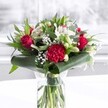 A lush, romantic bouquet in a clear cylindrical glass vase sits on a clean white surface, glowing softly as if near a bright Lea Bridge window on a spring morning. Inside the vase, neat green stems are visible through the fresh water, giving the arrangement a sense of honesty and life. Above, deep red roses and a large ruffled crimson carnation dominate the composition, their velvety petals unfolding in layers that suggest passion, commitment, and classic romance. Clustered between these bold blooms are creamy white alstroemeria and other pale blossoms, their slender petals marked with fine speckles that lend an almost painterly detail. Several white lily buds remain tightly closed, pointed upwards with quiet promise, hinting at flowers still to come and days of beauty ahead. Wisps of white gypsophila, or baby's breath, weave gently through the design like a light mist, softening the rich reds and adding a cloud-like texture that makes the bouquet feel full, airy, and generous. Around the base, broad, glossy dark green leaves are artfully wrapped and curled, forming a structured collar that frames the flowers and gives the piece its polished, contemporary shape. More narrow, eucalyptus-like foliage rises and fans out, introducing different greens and subtle movement, like leaves catching a Lea Bridge breeze along the River Lee. The lighting is even and natural, picking up every curve of petal and every vein in the foliage, so the colours remain rich but never harsh. This arrangement feels tailored for a meaningful moment-a romantic anniversary dinner in a Lea Bridge townhouse, a thoughtful birthday surprise for someone working from a local office, or a heartfelt Mother's Day delivery that brings warmth and gratitude into a family kitchen.