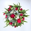 Viewed from directly above against a clean white backdrop, this lush hand-tied bouquet forms a generous circle of colour and texture, as if just set down on a florist's bench in Lea Bridge. At its centre, velvety crimson red roses, some still in tight bud and others beginning to unfurl, create a deep, romantic heart to the design. Around them, ruffled fuchsia and ruby red carnations add a playful, almost festive energy, their many frilled petals catching the light and giving the arrangement a sense of movement. Clusters of dainty white spray chrysanthemums, with their neat, daisy-like faces, punctuate the red tones with crisp brightness, helping the eye travel around the bouquet. Threaded delicately between these are clouds of tiny white gypsophila, or baby's breath, softening every edge and adding a dreamy, mist-like veil. Supporting this medley of blooms is a frame of rich greenery: broad, pointed dark green leaves, likely from lilies or alstroemeria, form a strong structure, while rounded, silvery eucalyptus leaves weave through with a cool blue-green note and a subtle, refreshing scent. The overall impression is of a full, thoughtfully crafted bouquet, fresh from a local Lea Bridge florist who understands both classic romance and modern style. It feels equally at home as a Valentine's Day surprise, an anniversary bouquet delivered to a townhouse off Lea Bridge Road, or a vibrant get well soon gift sent to brighten a friend's living room, carrying with it the warmth, vitality, and care of a hand-made floral gift.
