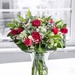 A lush, romantic bouquet of red roses, red carnations, white alstroemeria, and clouds of tiny white gypsophila stands proudly in a clear glass vase on a bright white table, bathed in gentle daylight from a large window. Each crimson rose shows off velvety petals in different stages of bloom, from tight buds promising tomorrow's beauty to open blossoms that curl softly at the edges, rich as velvet ribbon. Between them, full, ruffled carnations in the same deep red shade add texture and volume, creating the feeling of a traditional, heartfelt gift. Crisp white alstroemeria, some still in bud and others opening into trumpet-shaped flowers with pale green-tipped petals, weave between the reds, cooling the palette and drawing the eye deeper into the arrangement. Sprays of delicate gypsophila float like fine mist among the larger blooms, their tiny white stars softening every line. The bouquet is cradled by broad, glossy dark green leaves and threaded with more slender greenery, giving a sculpted, yet natural frame that feels freshly gathered. Through the vase, the neatly cut stems are visible, immersed in clear water that catches subtle reflections from the white surface below. The background outside the window is softly blurred and almost monochrome, like a quiet Lea Bridge street on a cloudy morning, so all attention rests on the vivid reds, whites, and greens in front. This design feels like a note of appreciation or love, ideal for birthdays, anniversaries, or simply sending a bright, uplifting message to someone in Lea Bridge without saying a word.