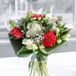 A contemporary mixed bouquet sits in a clear glass vase on a windowsill, its colours and textures glowing softly in the natural Lea Bridge light. At the heart of the arrangement, a coral-red rose and a rich crimson carnation provide a vivid focal point, their velvety petals layered and full, almost pulsing with warmth. Around them, clusters of dainty white chrysanthemums and tiny daisy-like blooms create a gentle halo, their crisp petals echoing the brightness of a cool morning along Lea Bridge Road. Several creamy white lilies, some still held in smooth green buds and others just beginning to open, rise gracefully through the design, adding a sense of poised anticipation and calm. The flowers are cradled by deep emerald foliage, the leaves folded and curved with a florist's precision to form wide arcs around the base, giving the bouquet a structured, modern silhouette. The glass vase is simple and clear, revealing clean stems and fresh water, and catching small glints of reflected daylight from the room. There is a feeling of fresh air and quiet celebration, as if the bouquet has just been delivered from a local florist to brighten a flat overlooking the River Lea. You can almost sense the gentle fragrance-a mix of lily sweetness, leafy green notes, and the soft spice of carnation-drifting through the space, making this arrangement perfect for marking a milestone or simply lifting the everyday mood in a Lea Bridge home.