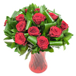 A lush, rounded bouquet of a dozen or more deep crimson roses fills the frame, each blossom opened to reveal velvety, tightly spiraled petals that catch the light with a subtle sheen. The red is intense-almost wine-dark in some shadows-and the soft, even studio lighting brings out the delicate gradations of pigment at the petal edges, lending the flowers a tactile, sumptuous quality. Dark green foliage is woven generously between the rose heads: glossy flat leaves that form a dense verdant backdrop and broader, sculpted leaves that have been artfully curled and rolled into elegant spirals. These architectural green elements-reminiscent of Aspidistra folds and Ruscus collars-add modern structure and cradle each bloom, their smooth surfaces contrasting with the rose petals' plush texture. The stems are gathered into a tall, slender translucent ruby-red glass vase whose subtle ribbed or twisted pattern creates vertical lines that catch and refract light, echoing the roses' passionate hue and adding a refined rhythm to the composition. Set against a crisp white background, the saturated reds and emerald greens pop with clean clarity, isolating the arrangement so every detail reads clearly: petal veining, glossy leaf surfaces, and the interplay of shadow and reflection on glass. There is an implied scent of classic roses-a gentle, familiar perfume-and a quiet elegance that makes this arrangement feel perfectly suited to anniversaries, Valentine's moments, or an intimate, romantic centerpiece. A florist's touch is evident in the balance of volume and negative space, a design that reads equally as a heartfelt gift or a sophisticated display for an evening table near Lea Bridge Road or a riverside dinner by the River Lea.