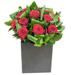 From where I stand, this arrangement reads like a study in modern romance: a compact cluster of deep crimson roses set against a chorus of glossy, emerald-green leaves that have been gently curled and folded to suggest movement. Each rose displays velvety petals that spiral outward with a plush, tactile richness; the light in the photograph is bright and steady, catching tiny highlights on the petal edges so the crimson appears almost luminous against the darker greens. The foliage is sculptural-broad, waxy leaves looped and layered to form a rhythmic backdrop that lifts and supports the roses without stealing the stage. All of this sits neatly within a matte black square vessel, its cube silhouette lending a contemporary, urban anchor that reads like a miniature sculpture on a coffee table or reception desk. Close up, you can almost sense the contrast between the roses' soft silkiness and the cool, firm texture of the foliage; there is a faint, clean scent of rose, crispened by green, almost tropical notes from the leaves. This bouquet feels designed for thoughtful declarations: a quietly dramatic anniversary gift, a romantic surprise delivered across Lea Bridge Road, or a polished statement piece for a city flat looking for warmth. As a local florist who often works around the River Lea and the marshes, I see how the arrangement's balance of bold colour and structured greenery fits both intimate dining tables and contemporary office spaces, promising to brighten a moment with deliberate, tactile beauty.